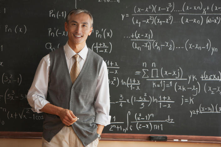 A teacher smiling in front of a chalkboard with hundreds of equations artfully written behind him.